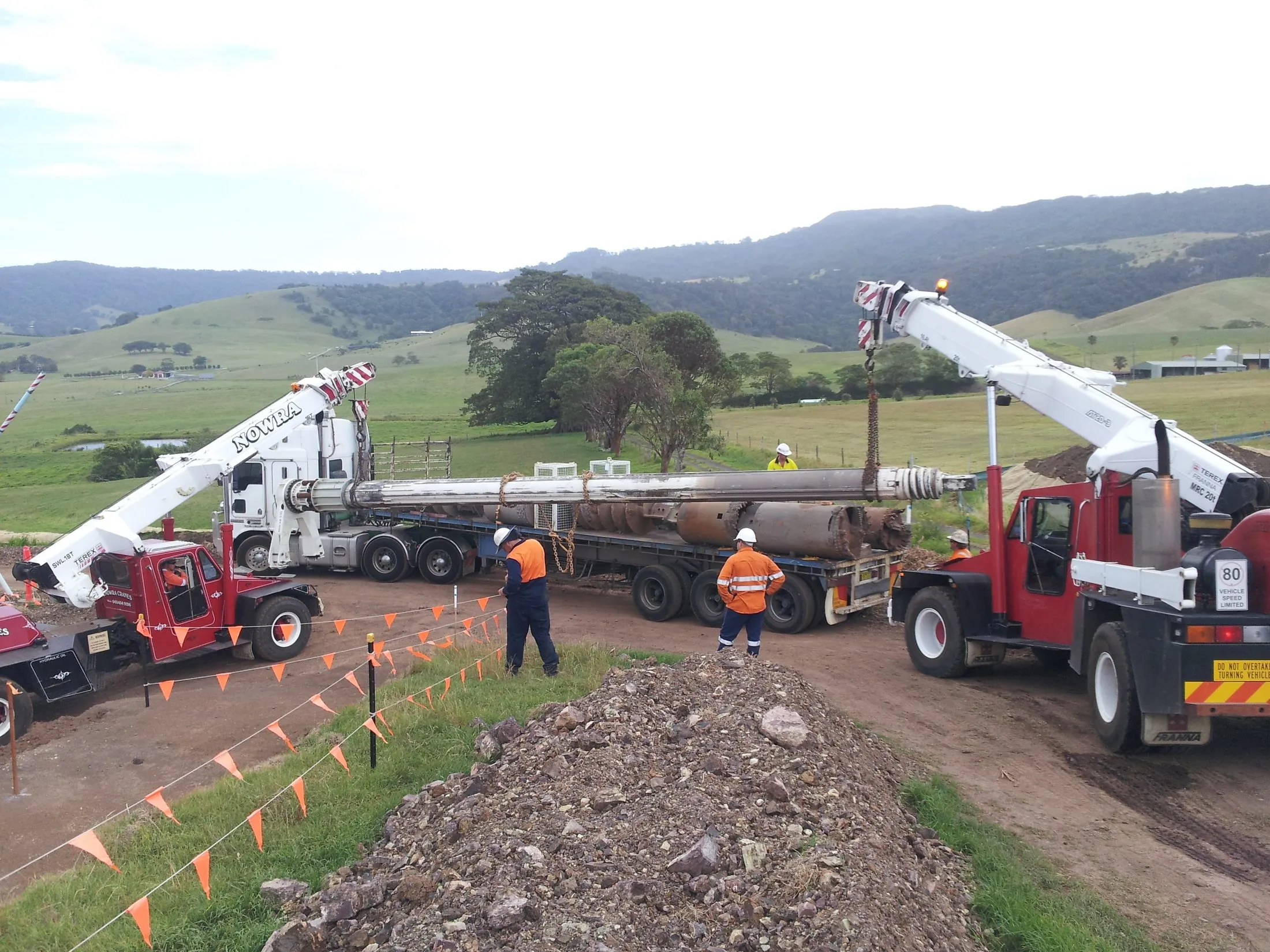 Two cranes lifting a long pipe on a farm